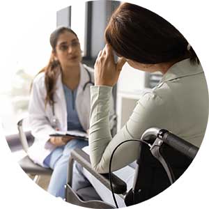 A woman sitting in a wheelchair holding her head and talking to a woman wearing scrubs and a white coat