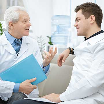 An older man in a white lab coat talks to a younger man in a white lab coat
