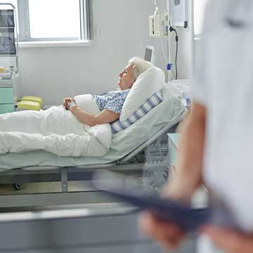 A woman in the background lays in a hospital bed with a health care worker in the foreground