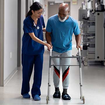 A person uses a walker alongside a person in blue scrubs